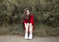 Portrait of young brunette young woman seats on grass, wearing red dress and white sneakers, looking at the side