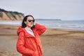 Portrait of a young brunette woman in sunglasses and a red jacket. Woman on a walk along the sandy shore of a large river on a spr