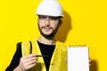 Portrait of young builder engineer with safety helmet and glasses, explains showing a pen on notebook with mockup on yellow. Royalty Free Stock Photo