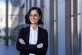 Portrait of a young business woman in glasses and a suit standing outside an office building with her arms crossed on Royalty Free Stock Photo