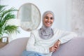 Portrait of young business woman of oriental origin in hijab sitting on sofa at home and looking at camera confidently