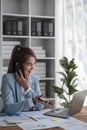 Portrait of young business woman with sitting in office in front of her laptop and talking on mobile phone hand at Royalty Free Stock Photo