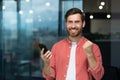 Portrait of a young businessman standing in the office, holding a mobile phone and happy showing a victory gesture with Royalty Free Stock Photo