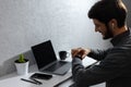 Portrait of young businessman verifies time on smartwatch, using wireless earphones. Laptop, coffee mug, cactus and smartphones.