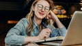 Portrait young businesswoman in fashionable glasses, sitting in cafe in front of computer and taking notes in notebook. Royalty Free Stock Photo