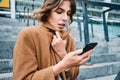 Portrait of young businesswoman fastening up coat from cold using cellphone outdoor