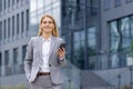 Portrait of a young businesswoman in a suit walking outside an office center, holding a phone and hand in her pocket Royalty Free Stock Photo