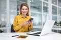 Portrait of a young businesswoman working in the office at a laptop, holding a mobile phone and smiling at the camera Royalty Free Stock Photo