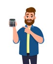 Portrait of young, casual dressed, bearded man showing or holding a digital calculator device in hand and pointing finger.