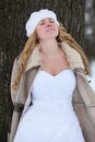 Portrait of young Caucasian bride in white wedding dress and fur coat leaning to a tree trunk with closed eyes Royalty Free Stock Photo