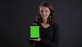 Portrait of young caucasian long-haired girl smilingly showing green screen of tablet to camera on black background.