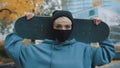 Portrait of young caucasian woman skater with protective face mask holding skateboard behind her head in the park in Royalty Free Stock Photo