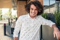 Portrait of young cheerful male model smiling with curly hair and freckles, wears trendy shirt, posing outdoor cafe and looking at Royalty Free Stock Photo