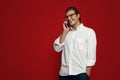 Portrait of a young cheerful man wearing white shirt and eyeglasses having phone call on red backdrop. Royalty Free Stock Photo