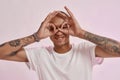 Portrait of young cheerful tattooed mixed race guy in white t shirt smiling at camera, making glasses gesture with his