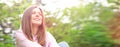 Portrait of a young cheerful woman sitting in the park on the green grass in a pink sweater hoodie.Wide image of a person at sun