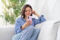Portrait of a young cheerful woman sitting on a sofa in the living room at home, laughing with her mobile phone, smartphone in her Royalty Free Stock Photo