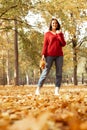 Portrait of young cheerful woman standing near trees among yellow leaves, holding bunch of maple leaves in forest park.