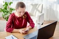 Portrait of young confident business woman sitting at wood desk with notepat and laptop computer in modern office and using mobile Royalty Free Stock Photo