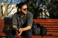 Portrait of young confident man sitting on wooden outdoor bench in the park, holding smartphone in hands. Wearing sunglasses. Royalty Free Stock Photo