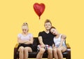 Portrait of young couple sitting on sofa with female friend feeling left out over yellow background