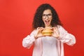 Portrait of young curly beautiful hungry woman eating burger. Isolated portrait of student with fast food over red background.
