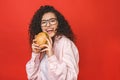 Portrait of young curly beautiful hungry woman eating burger. Isolated portrait of student with fast food over red background.