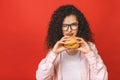 Portrait of young curly beautiful hungry woman eating burger. Isolated portrait of student with fast food over red background.