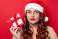 Portrait of a young curly girl with a red santa hat is holding a gift box with a red bow on a background of red wall Royalty Free Stock Photo