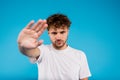 Portrait of young curly man in white t-shirt holds his hand showing stop sign on blue background Royalty Free Stock Photo