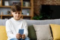 Portrait of young cute boy holding smartphone sitting at sofa. Teenager at home choosing favorite music, texting