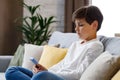 Portrait of young cute boy holding smartphone sitting at sofa. Teenager at home choosing favorite music, texting