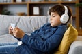 Portrait of young cute boy wearing earphones and holding smartphone sitting at sofa. Teenager at home texting messages