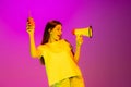 Portrait of young delightful girl posing, shouting in megaphone isolated over pink background in neon light