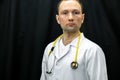 Portrait of a young doctor in a white coat on a black background. A doctor in blue gloves and with a medical stethoscope around Royalty Free Stock Photo