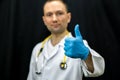Portrait of a young doctor in a white coat on a black background. A doctor wearing blue gloves and with a medical stethoscope Royalty Free Stock Photo