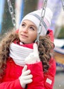 A portrait young dreamy girl in a red down-padded jacket and white hat sitting on a swing on the street in the park. Christmas Royalty Free Stock Photo