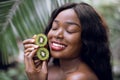 Portrait of young dreamy pleasant black woman with perfect smooth skin and natural nude make up, holding halfs of kiwi