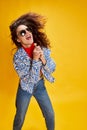 Portrait of young emotional girl posing in stylish casual clothes, singing in lollipop isolated over yellow background