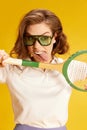 Portrait of young emotive young woman in sunglasses, posing with retro tennis racket against yellow studio background