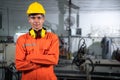 Portrait of young engineer standing and work in the robot factory. Wearing orange uniform, safety helmet and earmuffs crossed arms