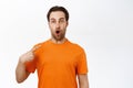 Portrait of young enthusiastic man looking surprised, pointing at himself, wearing orange t-shirt, white studio Royalty Free Stock Photo