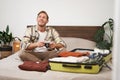 Portrait of young excited man, sits on bed with suitcase, going on holiday, holding digital camera, going through photo Royalty Free Stock Photo