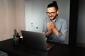 Portrait of young excited man working home at laptop, wearing eyeglasses and shirt, sitting at desk, background of grey wall Royalty Free Stock Photo