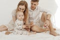 Portrait of young family with cherubic infant baby, with blue eyes holding wooden natural teether sitting on white bed.