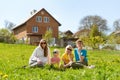 Portrait of young family sitting in the grass against two-story house. Mom or nanny sitting with children in meadow surrounded by