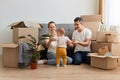 Portrait of young family unpacking moving box at their new apartment, sitting on floor near sofa and playing with their infant kid Royalty Free Stock Photo