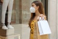 Portrait of young fashion woman with protective mask and shopping bags looking through shop window