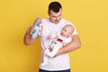 Portrait of young father holding his newborn baby in hands, playing and showing soft toy to calm down kid, guy wearing white Royalty Free Stock Photo