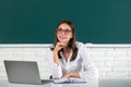 Portrait of young female college student in glasses studying in classroom on laptop in class with blackboard background. Royalty Free Stock Photo